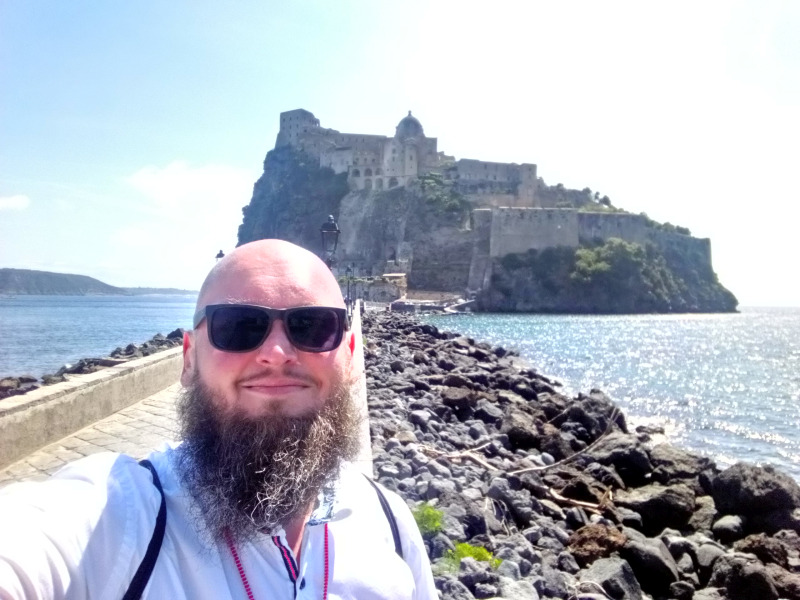 Markus Patho vor dem Castello Aragonese Ein Mann mit Bart und Sonnenbrille macht ein Selfie vor einer historischen Festungsanlage auf einem Hügel am Meer. Sonnenlicht reflektiert auf dem Wasser und die Felsen am Ufer sind deutlich sichtbar.