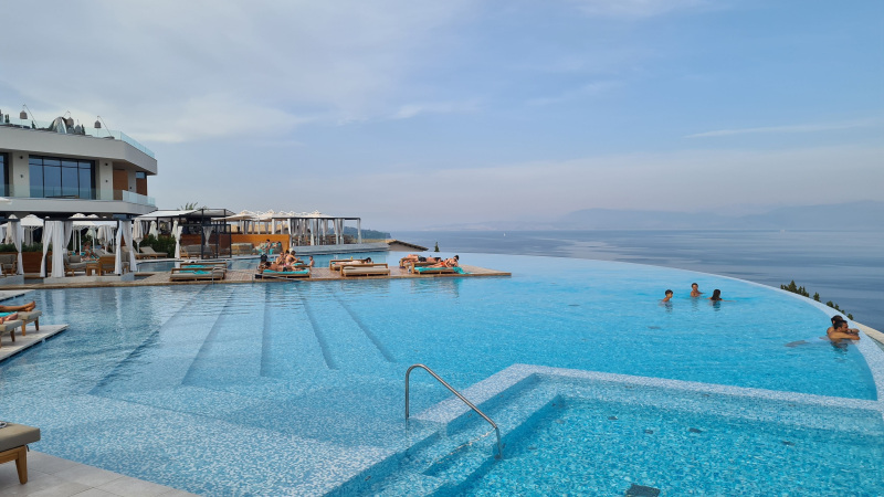 Infinity Pool Ein luxuriöses Infinity-Pool mit klarem blauen Wasser und Besuchern, die sich entspannen und schwimmen. Der Pool überblickt eine malerische Küstenlinie. Im Hintergrund ist ein modernes Gebäude mit Sonnenschirmen und Liegestühlen zu sehen. Himmel und Meer verschmelzen am Horizont.
