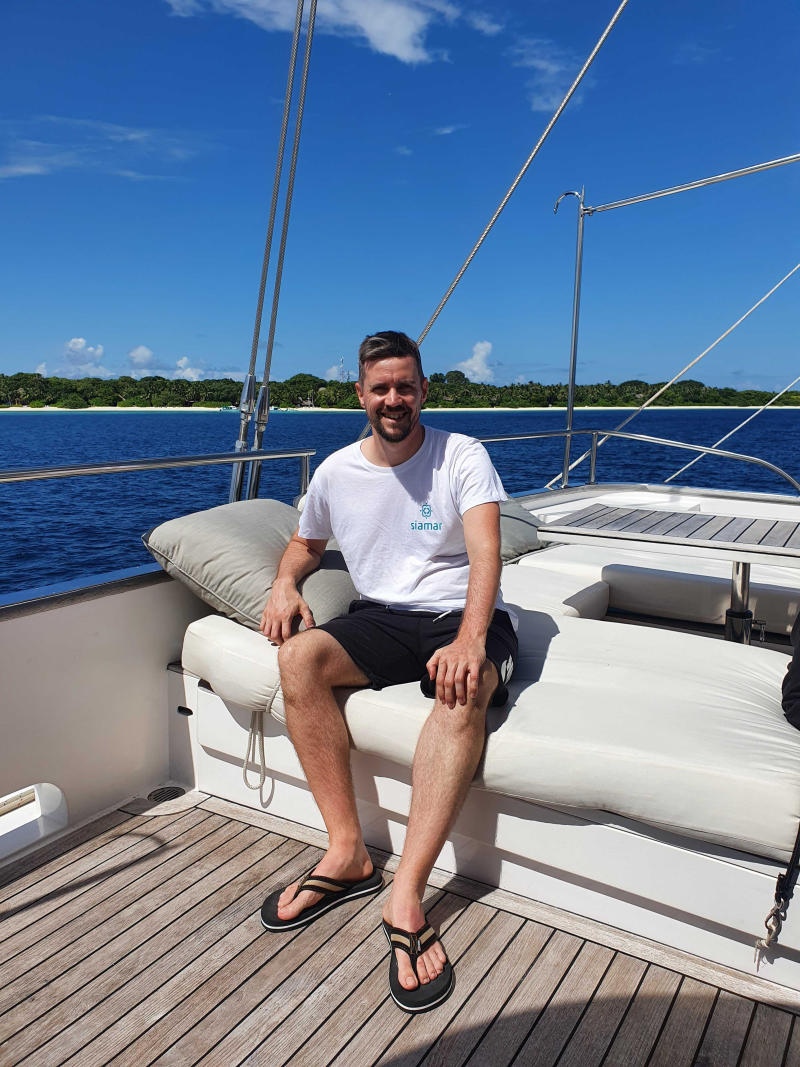 Christoph Busch auf der Yacht Ein lächelnder Mann sitzt entspannt auf einer cremefarbenen Sitzbank an Deck einer Yacht. Er trägt ein weißes T-Shirt, schwarze Shorts und Flip-Flops. Im Hintergrund ist bei strahlendem Sonnenschein ein ruhiges, blaues Meer mit einer grünen Insel zu sehen.