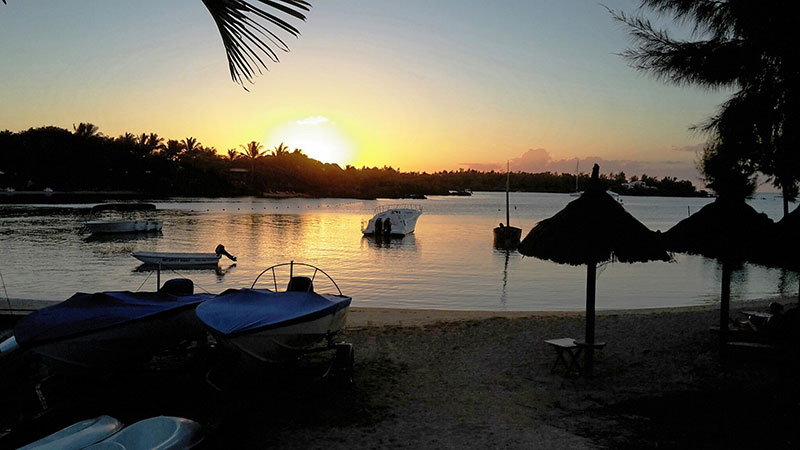 Sonnenuntergang auf Mauritius Bild eines ruhigen Strandes bei Sonnenuntergang mit kleinen Booten im Wasser, überdachten Liegen am Sandstrand und Palmen, die einen malerischen Rahmen für den orange-gelb gefärbten Himmel bieten.