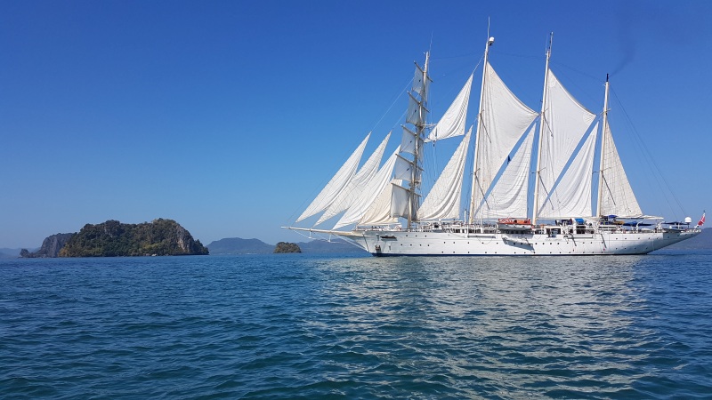 Die Star Clipper auf hoher See Ein großes Segelschiff mit weißen Segeln auf dem blauen Meer mit einer kleinen Insel im Hintergrund und einem klaren blauen Himmel.