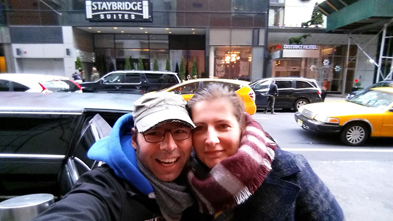 Silvestertrip nach New York – ein Traum Ein fröhliches Pärchen macht ein Selfie auf der Straße vor einem Hotel mit der Aufschrift "Staybridge Suites". Im Hintergrund sind Autos und ein klassisches gelbes Taxi zu sehen.
