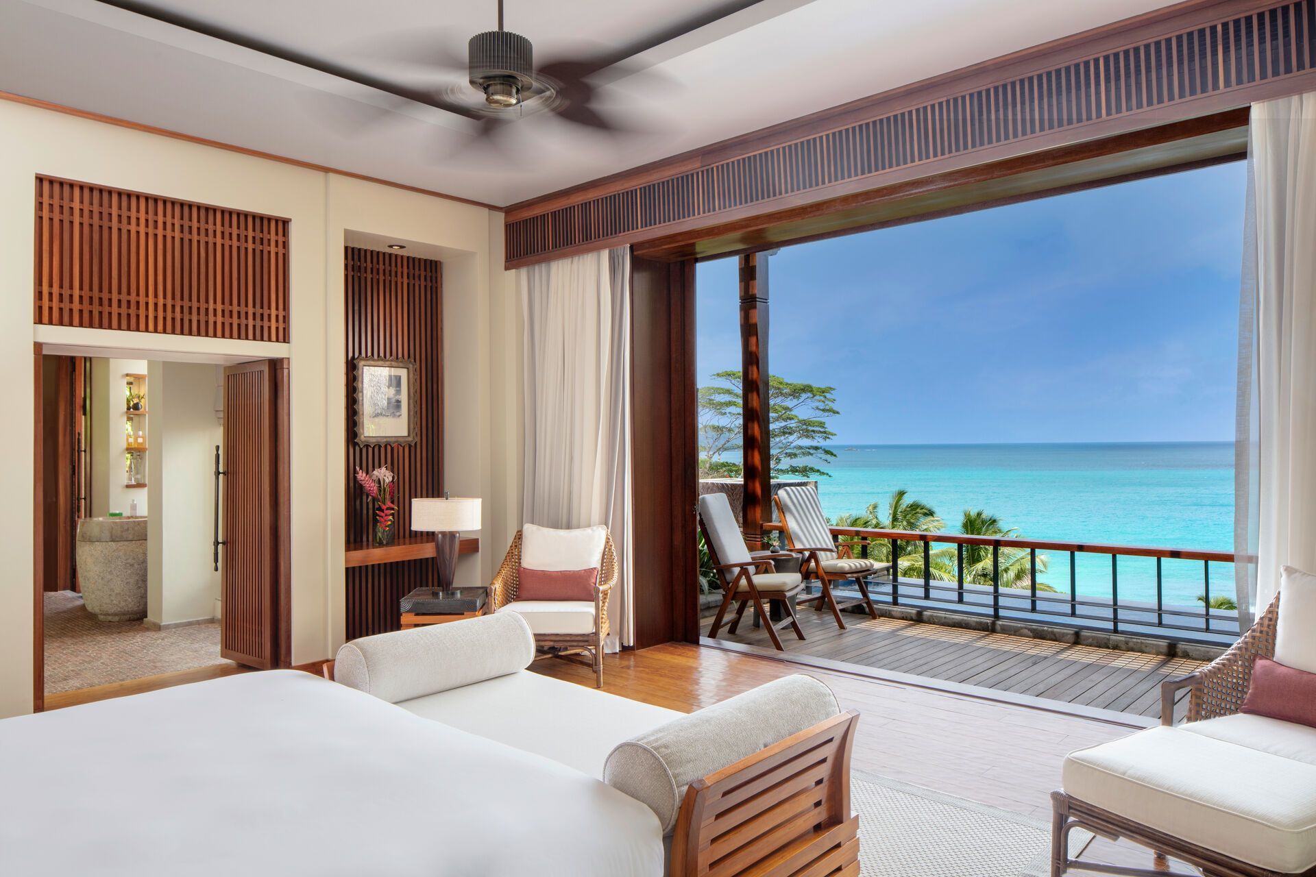 Ein luxuriöses Schlafzimmer mit großen Fenstern, die auf einen Balkon mit Meerblick führen. Das Zimmer ist elegant eingerichtet mit einem komfortablen Doppelbett, Sesseln und einer geschmackvollen Dekoration. Draußen strahlt das türkisblaue Meer und ergänzt die tropische Atmosphäre.