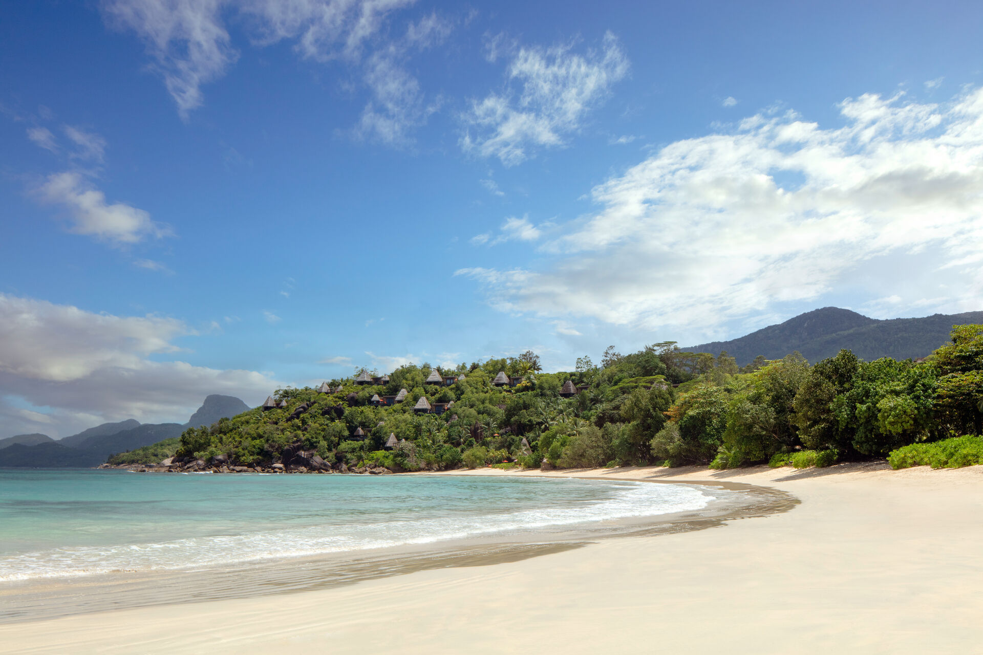 Blick auf einen idyllischen Strand mit weißem Sand und kristallklarem Wasser vor einer grünen, bewaldeten Hügellandschaft unter einem blauen Himmel mit wenigen Wolken. Am Hang sind Dächer von Verstecken zu erkennen, die zwischen den üppigen Bäumen hervorlugen.