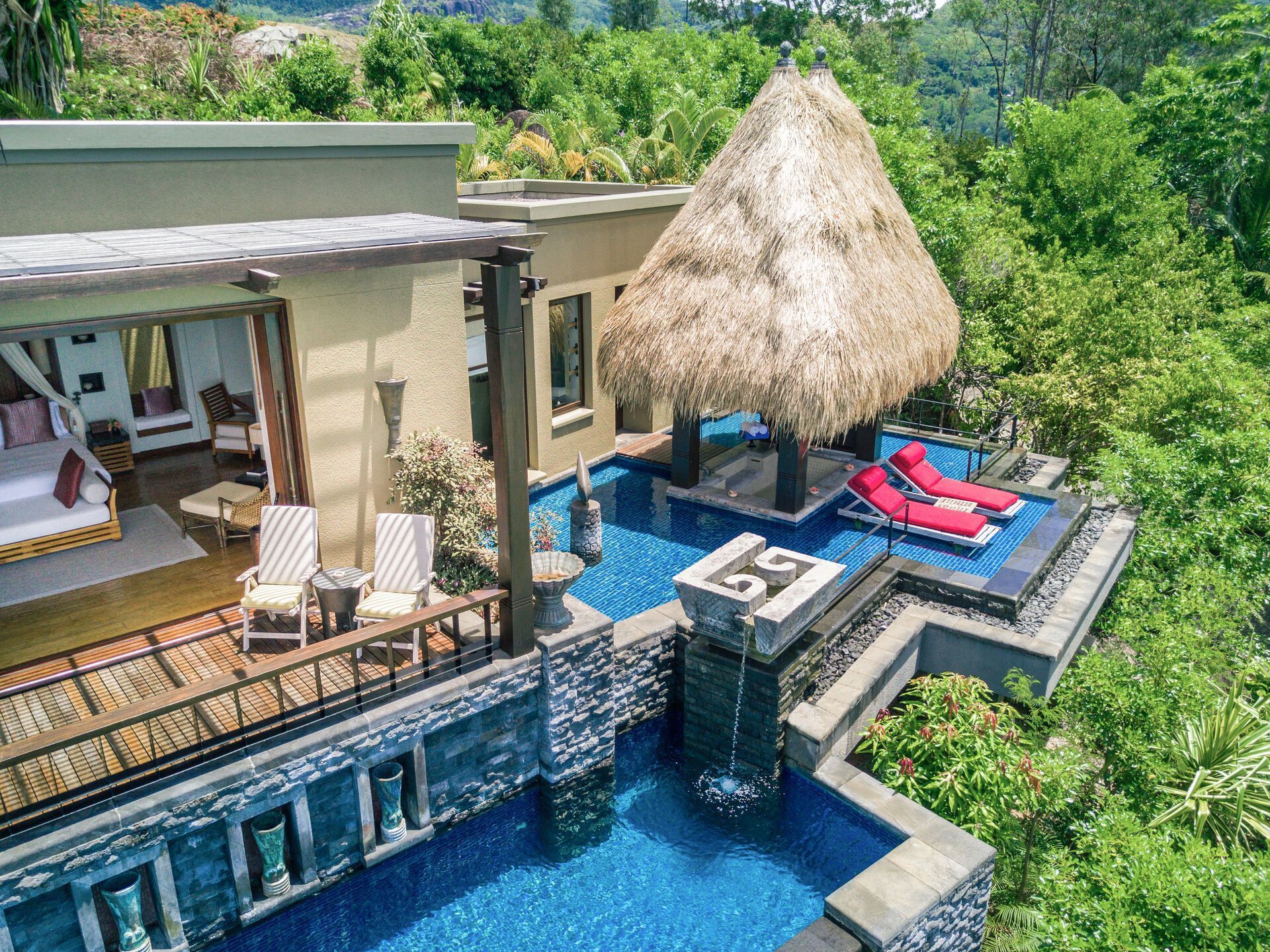 Luftaufnahme eines luxuriösen Ferienresorts mit strohgedecktem Pavillon, privatem Poolbereich mit Liegestühlen und Wasserfällen, umgeben von üppiger Vegetation.