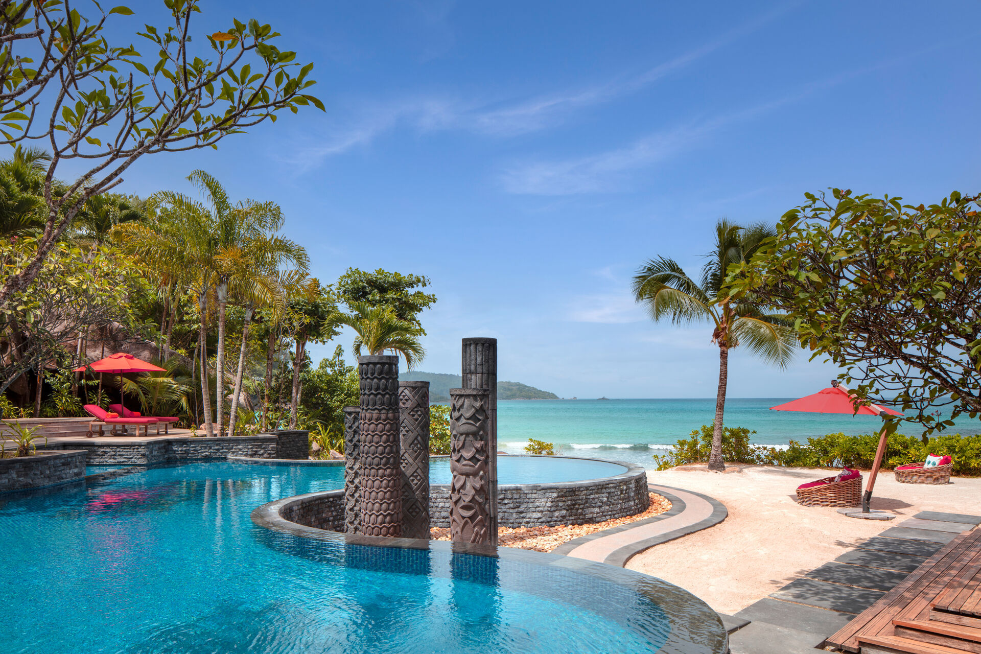 Ein luxuriöses Resort-Schwimmbad mit Blick auf das Meer, umgeben von tropischer Vegetation mit roten Sonnenschirmen und Liegestühlen.
