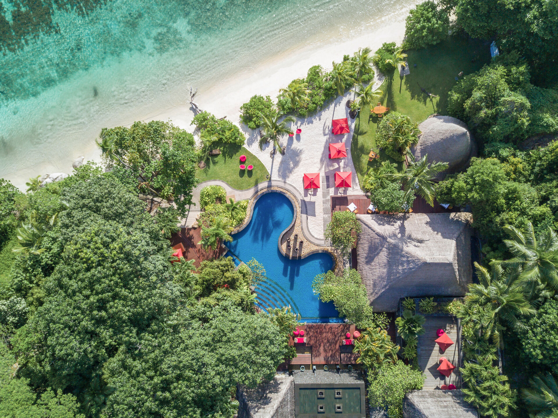 Luftaufnahme Luftaufnahme eines luxuriösen Resortkomplexes in tropischer Umgebung mit einem blauen Swimmingpool, strohgedeckten Pavillons und Liegestühlen unter roten Sonnenschirmen am Rand eines weißen Sandstrandes.