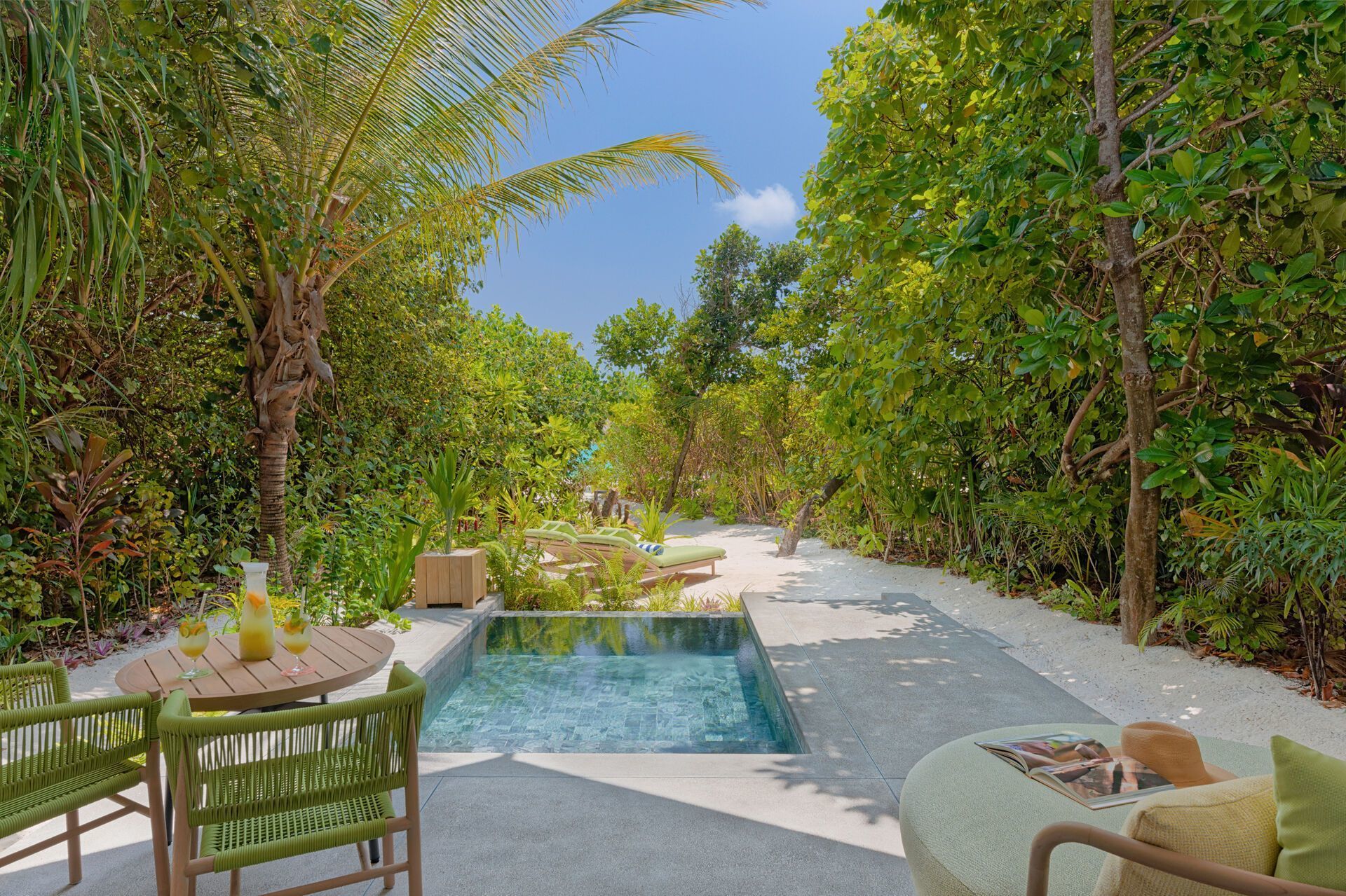 Einladende Freiluft-Lounge mit einem kleinen Pool umgeben von tropischem Grün, ausgestattet mit komfortablen grünen Stühlen, einem runden Tisch mit Erfrischungsgetränken und einer entspannenden Liegefläche im Hintergrund.