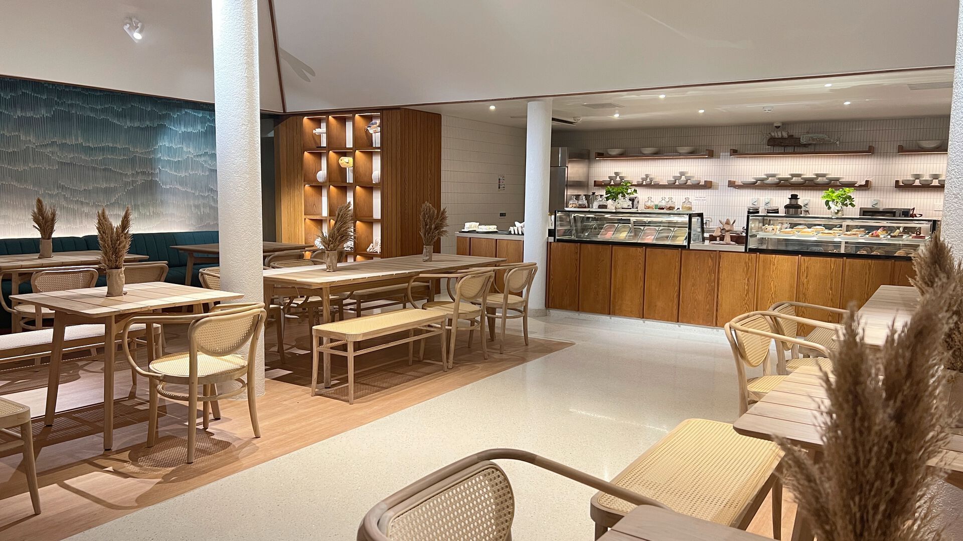 Alt-Text: Eine moderne Café-Inneneinrichtung mit hellen Holzmöbeln, bequemer Sitzbank an der Wand und dekorativen Getreideähren auf den Tischen. Eine Theke mit Kuchenauswahl befindet sich im Hintergrund rechts, und links sind Regale mit Tassen und Dekorationsgegenständen zu sehen.