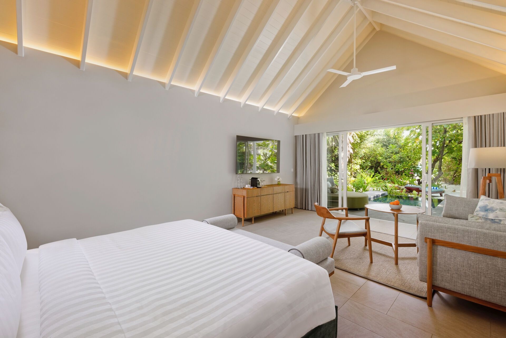 Ein modern eingerichtetes Hotelzimmer mit einem großen Bett, sauberer weißer Bettwäsche und hellen Wänden. Die abgeschrägte Decke mit versteckter Beleuchtung verleiht dem Raum eine offene und einladende Atmosphäre. Vor dem Bett befindet sich eine Sitzgruppe mit zwei stilvollen Stühlen und einem kleinen Tisch, die auf eine große Fensterfront mit Blick auf eine üppige, grüne Gartenanlage ausgerichtet ist. Am linken Rand des Bildes ist ein Holzmöbel mit Kaffeemaschine und Fernseher zu sehen. Der Raum ist durch seine Kombination von Komfort und modernem Design charakterisiert.