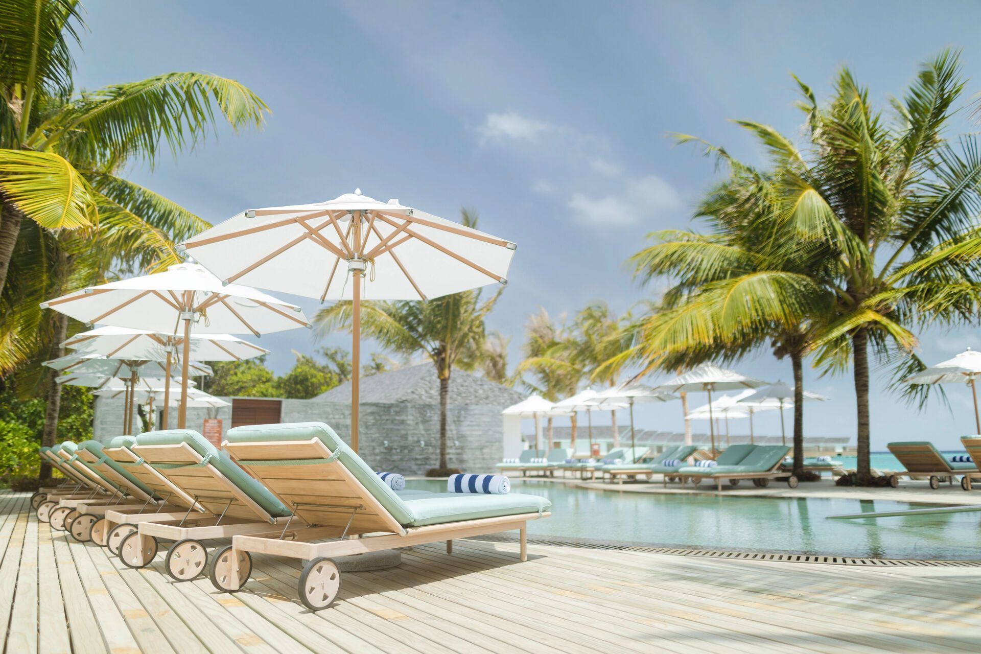 Ein luxuriöser Poolbereich in einem Resort mit Sonnenliegen unter geöffneten Sonnenschirmen, umgeben von Palmen und klarem Himmel, vermittelt eine entspannte Urlaubsatmosphäre.