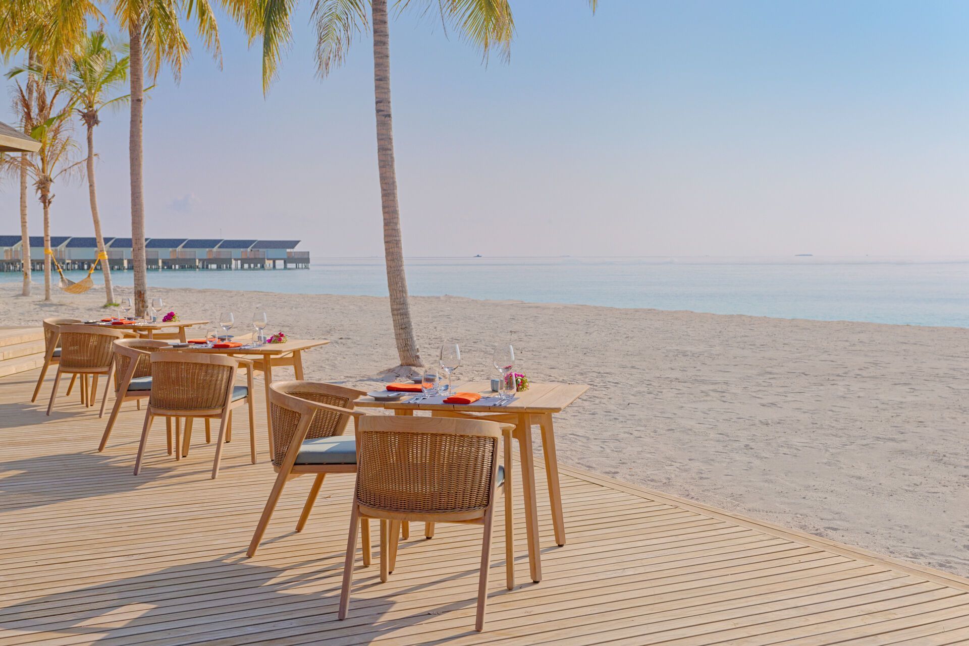 Am Strand Alternativtext: Ein idyllisches Strandrestaurant mit gedeckten Tischen und Stühlen aus Rattan auf einem Holzdeck, mit einem Blick auf ruhiges, türkisfarbenes Meer und über dem Wasser gebauten Bungalows in der Ferne. Palmen werfen leichte Schatten auf das Szenario, das Entspannung und Urlaubsgenuss suggeriert.