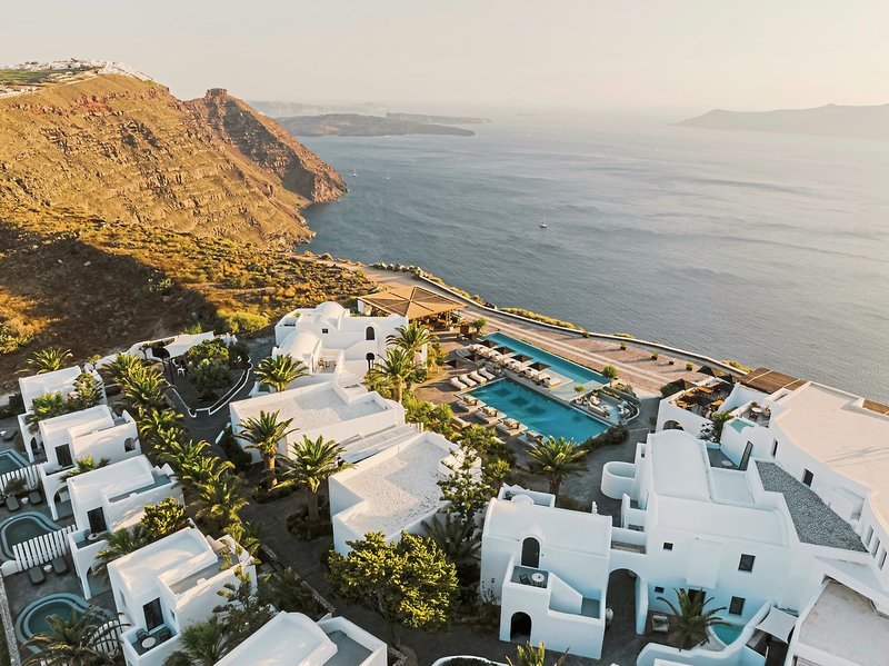 Lage auf der Klippe Blick auf eine Reihe weiß getünchter Gebäude im Kykladen-Stil mit blauen Pools und Palmen, die sich auf einer Klippe über dem ruhigen, blauen Meer der Ägäis befinden. Im Hintergrund ist eine hügelige Landschaft zu erkennen, die sanft ins Meer übergeht. Die Szenerie wird von warmem, goldenem Licht der tief stehenden Sonne beleuchtet.