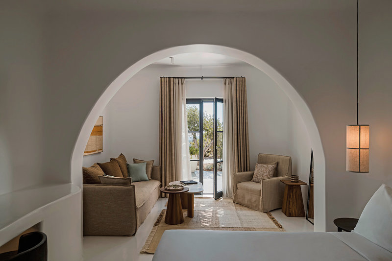 Innenarchitektur Ein elegantes, modern eingerichtetes Wohnzimmer mit geschwungenen weißen Wänden und hohen Decken. Im Zentrum steht ein beigefarbenes Sofa mit Kissen, gegenüber einem Sessel und einem kleinen Holztisch. Links befindet sich eine eingebaute Sitznische. Natürliches Licht fällt durch die geöffnete Doppeltür ein, welche auf eine Terrasse mit Blick auf die Vegetation führt. Rechts hängt eine stilvolle, quaderförmige Stehlampe an der Wand. Der Raum vermittelt eine ruhige und gemütliche Atmosphäre.