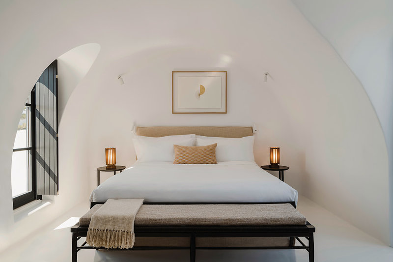 Alt-Text: Modern gestaltetes Schlafzimmer mit einer klaren, minimalistischen Einrichtung. Ein großes Doppelbett mit weißer Bettwäsche steht im Zentrum des Raumes, flankiert von zwei Nachttischen mit Leuchten. Über dem Bett hängt ein abstraktes Kunstwerk. Die Wände sind weiß und weisen eine geschwungene Architektur auf, wobei ein hohes, bogiges Fenster mit schwarzem Rahmen auf der linken Seite natürliche Beleuchtung bietet.