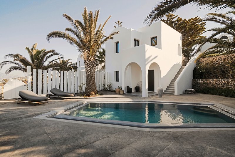 Außenanlage Eine weiße, kubisch gestaltete Villa mit flachem Dach und abgerundeten Ecken unter einem klaren Himmel. Vor dem Haus befinden sich ein nicht überdachter Pool in unregelmäßiger Form und zwei moderne Sonnenliegen. Umliegend sind Palmen und eine weiße, vertikale Holzumzäunung zu sehen, hinter der ein weiterer Gebäudekomplex teilweise sichtbar ist.