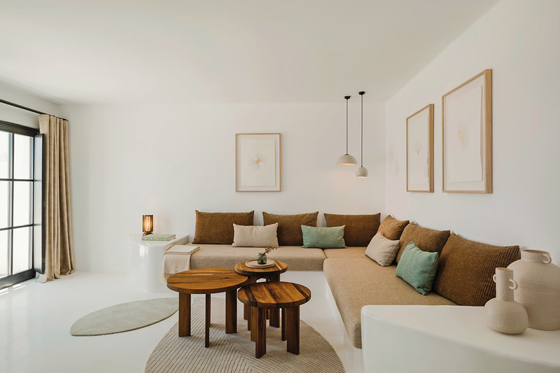 Ein helles, modernes Wohnzimmer in neutralen Farbtönen mit einem großen, beigen Ecksofa, das mit braunen und grünen Kissen ausgestattet ist. Runde Holztische stehen in der Mitte vor dem Sofa, und auf der linken Seite ist eine weiße Konsole mit einer leuchtenden Tischlampe zu sehen. An der Wand hängen drei gerahmte Kunstwerke mit abstrakten Motiven. Ein heller Teppich bedeckt teilweise den Boden, und die Szene erhält natürliches Licht durch eine große Fenstertür rechts im Bild.
