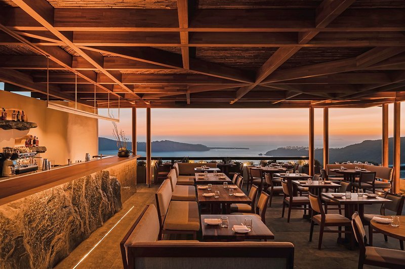 Ein elegantes Restaurant mit modernem Interieur bei Sonnenuntergang, das einen atemberaubenden Blick auf das Meer und eine Bergkette am Horizont bietet. Die Tische sind nett gedeckt und bereit für die Gäste. Warme Beleuchtung und Holzakzente sorgen für eine einladende Atmosphäre.