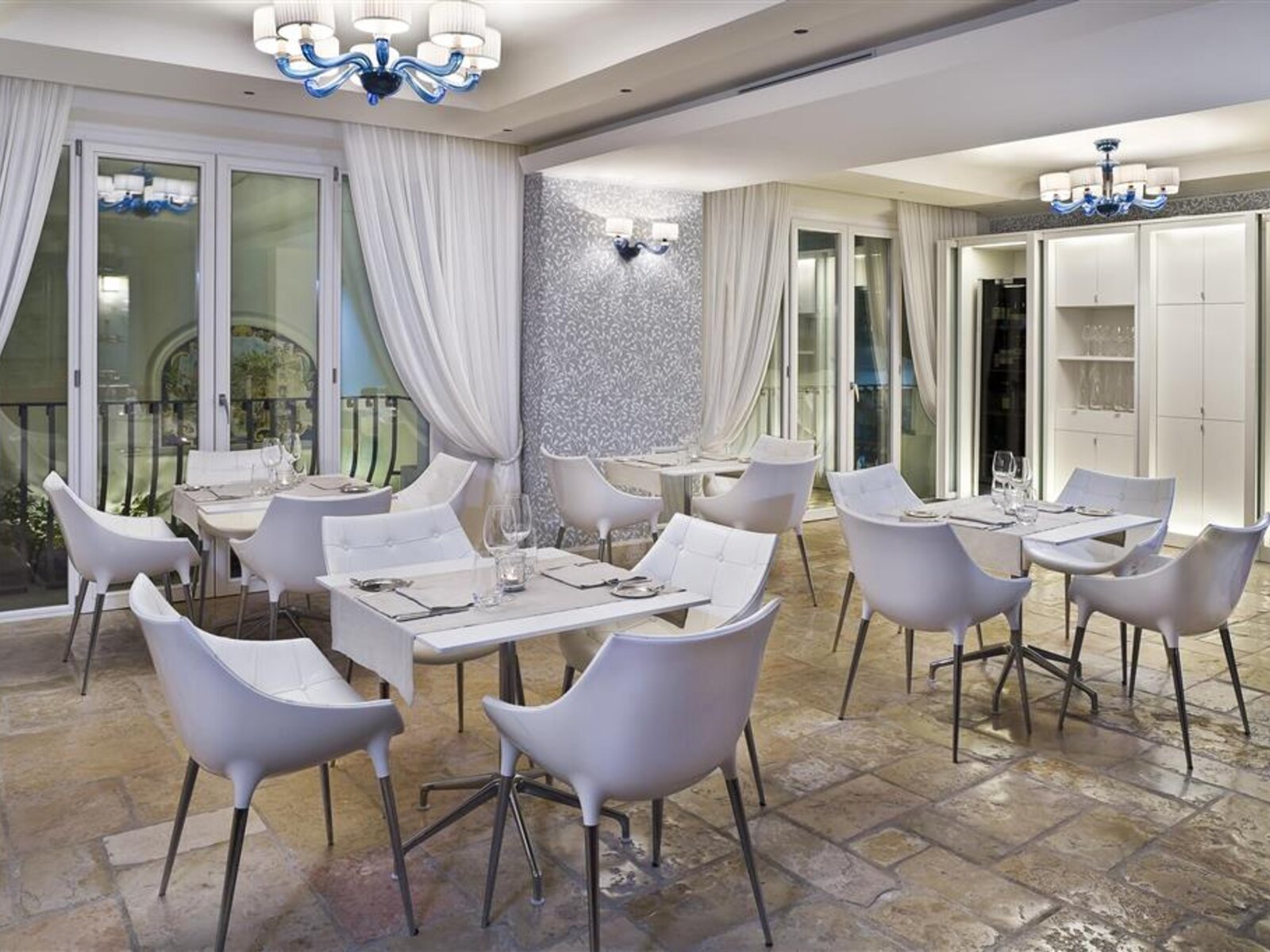 Alternativtext: Ein elegantes Restaurantinterieur mit modernen weißen Stühlen und Tischen, die für das Essen vorbereitet sind, auf einem rustikalen Steinboden. Im Hintergrund sind transparente Vorhänge vor Glastüren, die auf eine Außenterrasse führen, und an der Decke hängt eine Designerglasleuchte in Blautönen.