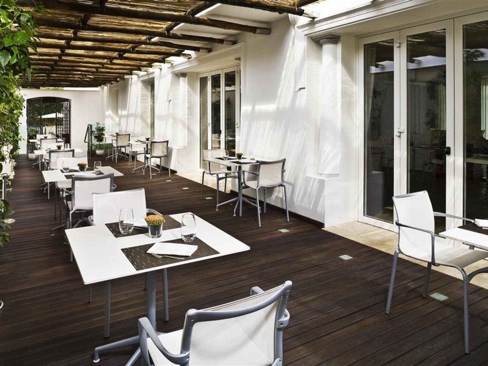 Restaurantterrasse des Hotel Villa Blu Capri Ein leerer Restaurantterrasse mit dunklem Holzboden und weißen Tischen sowie Stühlen unter einem rustikalen Holzdach. Weißer Vorhänge flattern im leichten Wind und Glasfronten führen ins Gebäudeinnere.