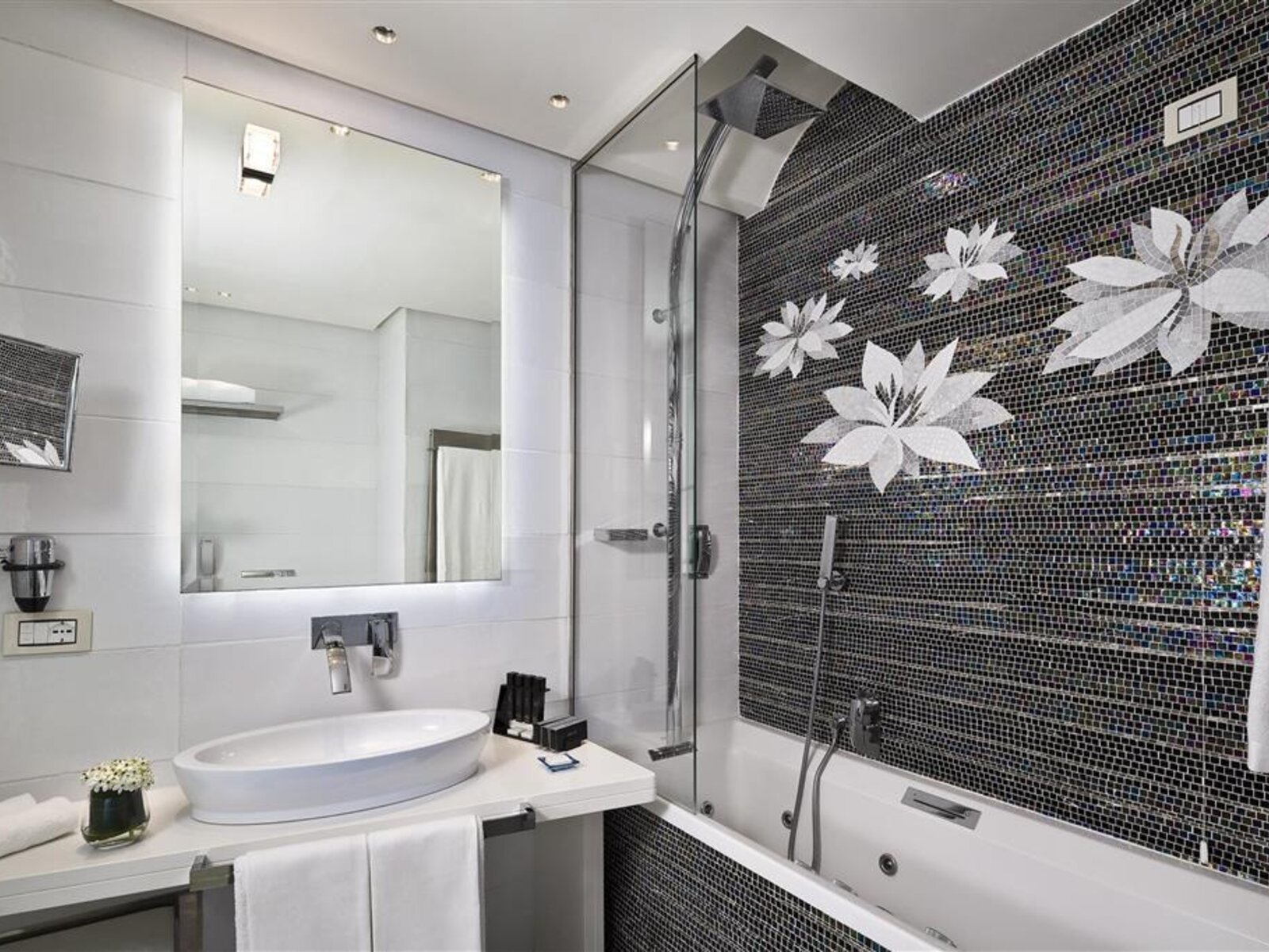Ein modern gestaltetes Badezimmer mit einer weißen Waschschüssel auf einer weißen Arbeitsplatte, großem Spiegel darüber und einer klaren Glastür, die eine Duschkabine mit schwarzem Mosaikfliesen und weißen Blumenmustern abschließt.