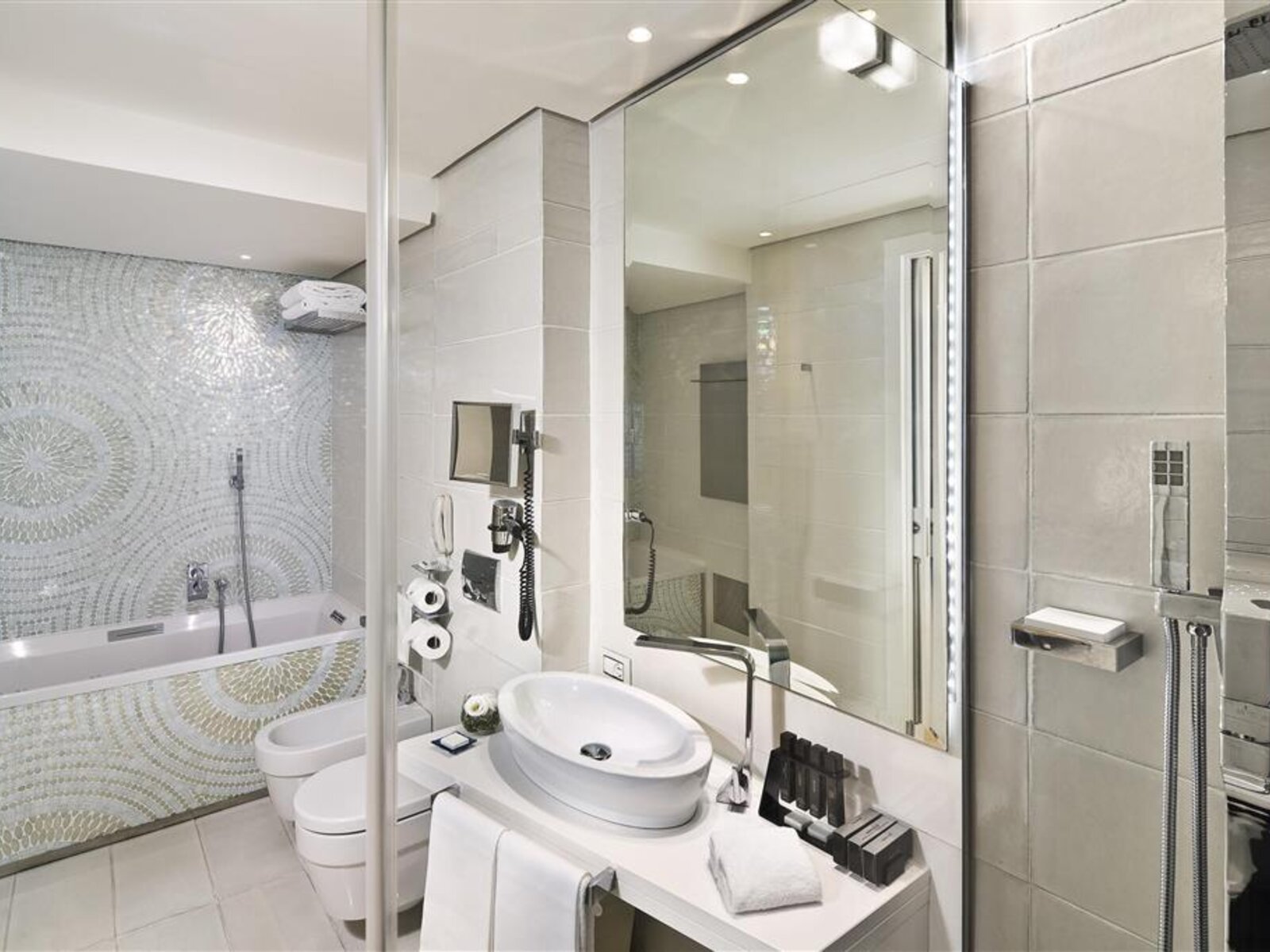 Ein modernes Badezimmer mit einer Mischung aus weißen und hellgrauen Tönen, ausgestattet mit einer Badewanne, einer separaten Duschkabine, einem wandmontierten WC und einem einzelnen Waschbecken. An der Wand befindet sich ein auffälliges Mosaikmuster, und das Bad verfügt über eine Vielzahl von Pflegeprodukten sowie Handtücher, die bereitliegen.