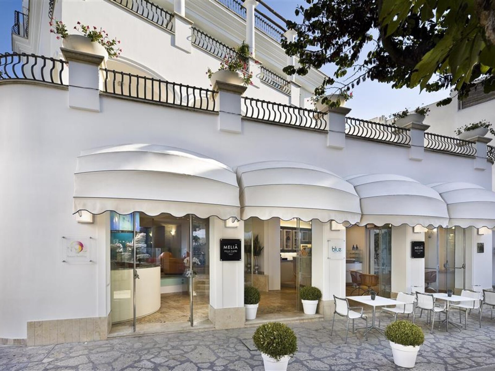 Elegantes Hotelfassade im traditionellen Stil mit einer Außenterrasse und ordentlichen Tischgruppen. Über der Eingangstür ein Schild mit der Aufschrift "MELIA". Über der weißen Fassade sieht man Balkone mit schmiedeeisernen Geländern und Blumenkästen.