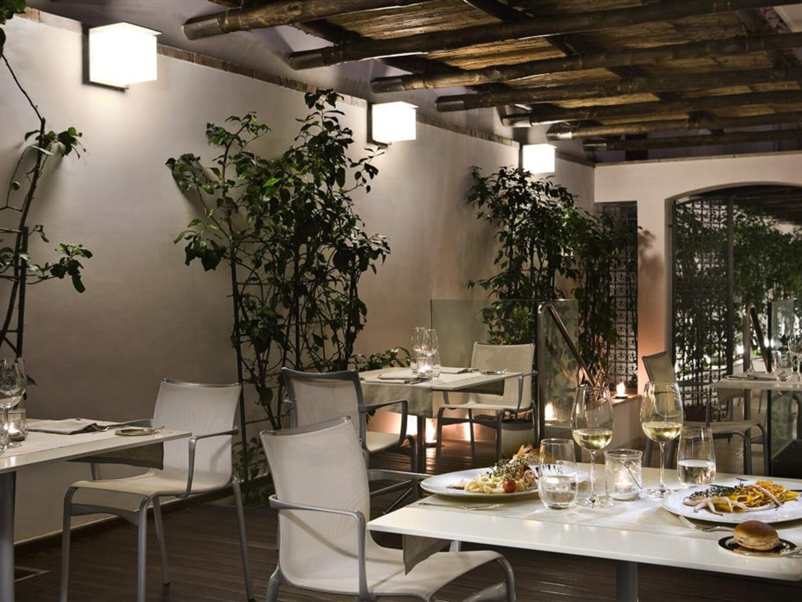 Restaurant Ein stilvolles Restaurantinterieur mit modernen weißen Stühlen und Tischen, auf denen Teller mit Essen und Weingläser stehen. Rustikale Holzbalken zieren die Decke und an der Wand wachsen hohe Grünpflanzen, die eine gemütliche Atmosphäre erzeugen. Warmes Licht strahlt von Wandleuchten aus und sorgt für eine einladende Stimmung.