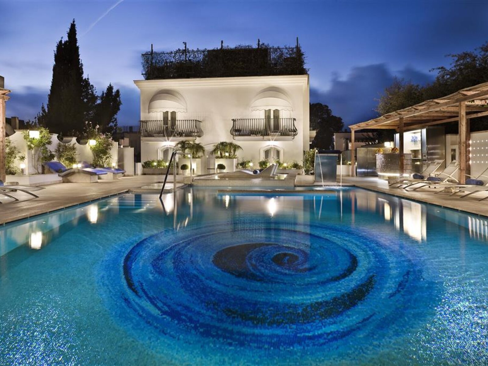 Abendstimmung im Hotel Villa Blu Capri Ein luxuriöses Pool-Setting während der Dämmerung mit beleuchteten Liegestühlen, einem eleganten weißen Gebäude im Hintergrund und einem kunstvollen spiralförmigen Muster auf dem Wasser.