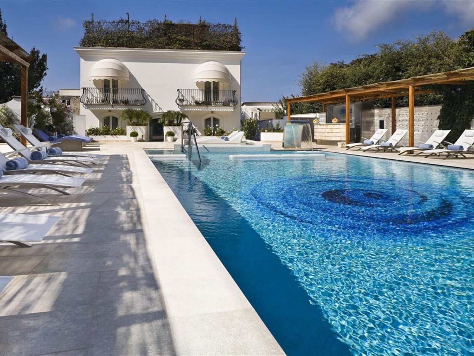 Luxuriöser Außenpoolbereich einer eleganten Villa mit blauem Wasser und einem dekorativen Mosaik am Boden des Pools. Weiße Liegestühle stehen am Poolrand, daneben ist ein bedachter Ruhebereich. Im Hintergrund befindet sich die weiße Fassade des zweistöckigen Gebäudes mit Balkonen und rankenden Pflanzen.