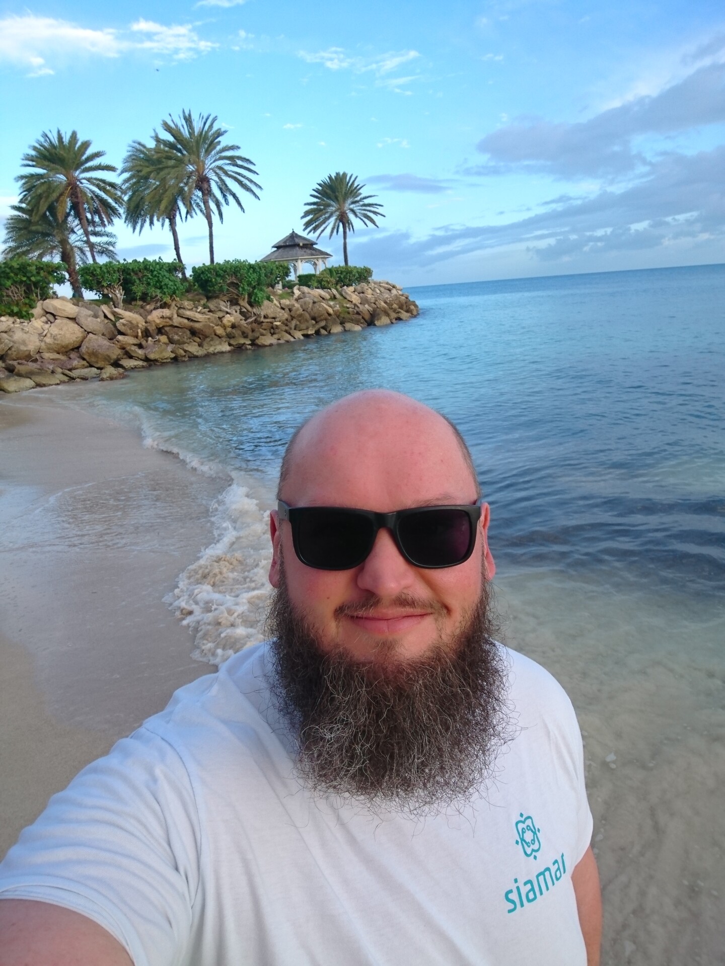 Markus Patho am Strand von Antigua Ein Mann mit Vollbart und Sonnenbrille macht ein Selfie am Strand. Im Hintergrund sind Palmen, ein Pavillon auf einem Steinwall und ein ruhiges Meer unter einem leicht bewölkten Himmel zu sehen.