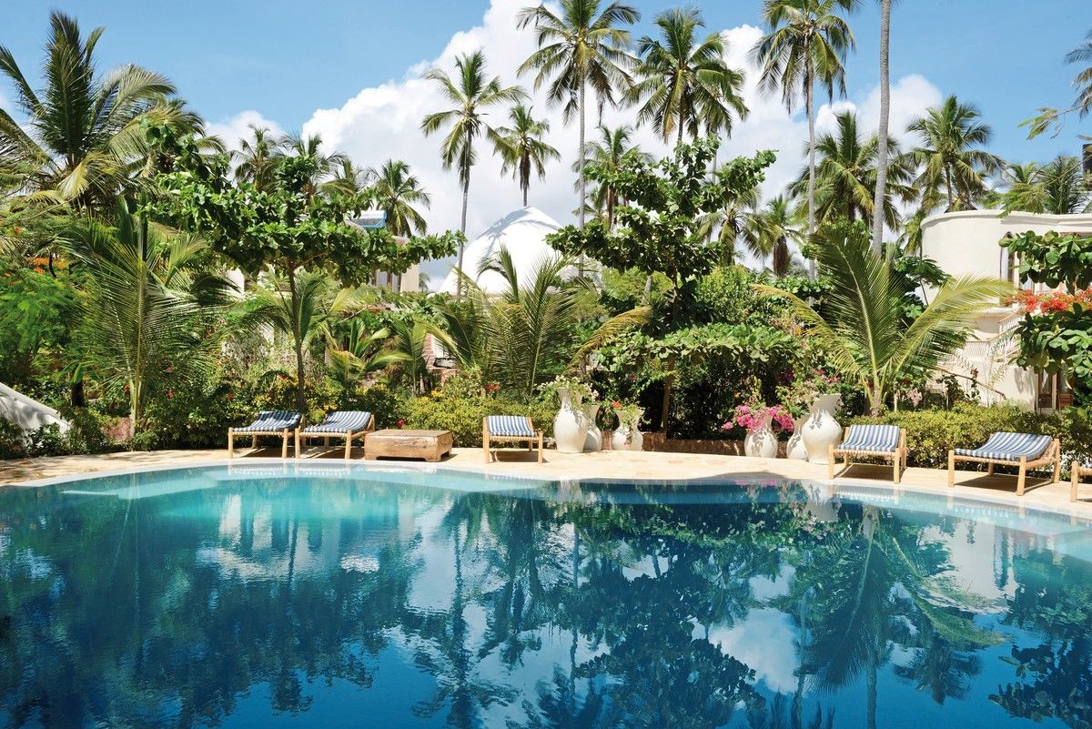Ein luxuriöses Resort-Schwimmbad umgeben von hohen Palmen und üppiger tropischer Vegetation unter einem klaren, blauen Himmel. Liegestühle stehen am Rande des Pools bereit für Entspannung.