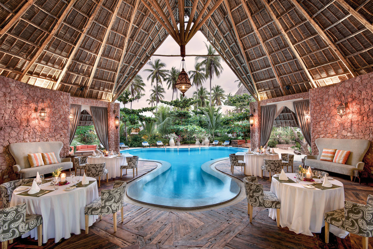 Ein luxuriöses Open-Air-Restaurant mit einem reetgedeckten Dach, das eine warme und einladende Atmosphäre ausstrahlt. Im Vordergrund sind elegant eingedeckte Tische mit weißen Tischdecken und geschmackvoller Tischdekoration zu sehen. Ein ansprechender Swimmingpool im Zentrum bildet das Herzstück des Raumes, umgeben von üppigem Grün und Steinwänden, die einen natürlichen Kontrast zum blauen Wasser bieten.