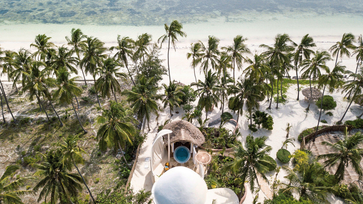 Luftaufnahme eines tropischen Strandresorts mit strohgedeckten Hütten umgeben von zahlreichen schlanken Kokospalmen. Im Vordergrund sind Wege zu sehen, die sich durch den weißen Sand schlängeln, und ein kristallklares Meer liegt im Hintergrund.