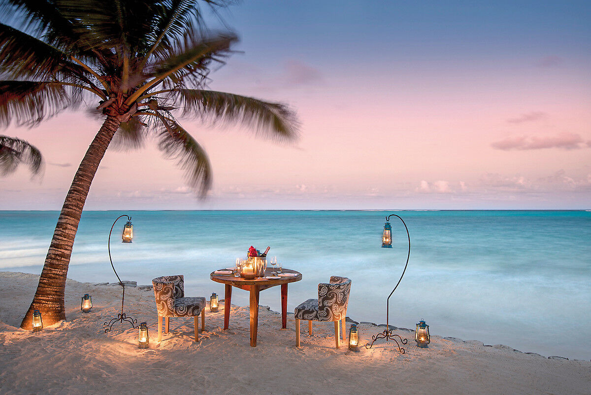 Ein romantisch gedeckter Tisch für zwei Personen am Strand bei Sonnenuntergang, mit einer sich neigenden Palme und Laternen, die für eine stimmungsvolle Beleuchtung sorgen. Der Ozean ist ruhig und der Himmel in Pastelltönen von Rosa und Blau gefärbt.