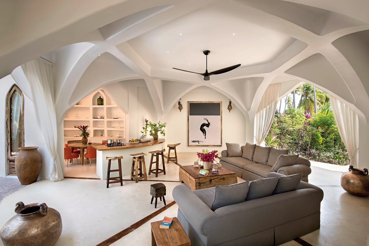Ein stilvoll eingerichtetes Wohnzimmer mit gewölbter Deckenarchitektur und modernen organischen Formen. Im Vordergrund steht ein großes graues Ecksofa rechts, links befindet sich ein rustikaler Holztisch mit Hockern. An der Wand hängt ein Kunstwerk mit einem schwarzen Vogel auf weißem Hintergrund. Ein großes Bogenfenster öffnet den Blick auf eine üppige Gartenlandschaft. Das Ambiente vermittelt eine warme und einladende Atmosphäre.