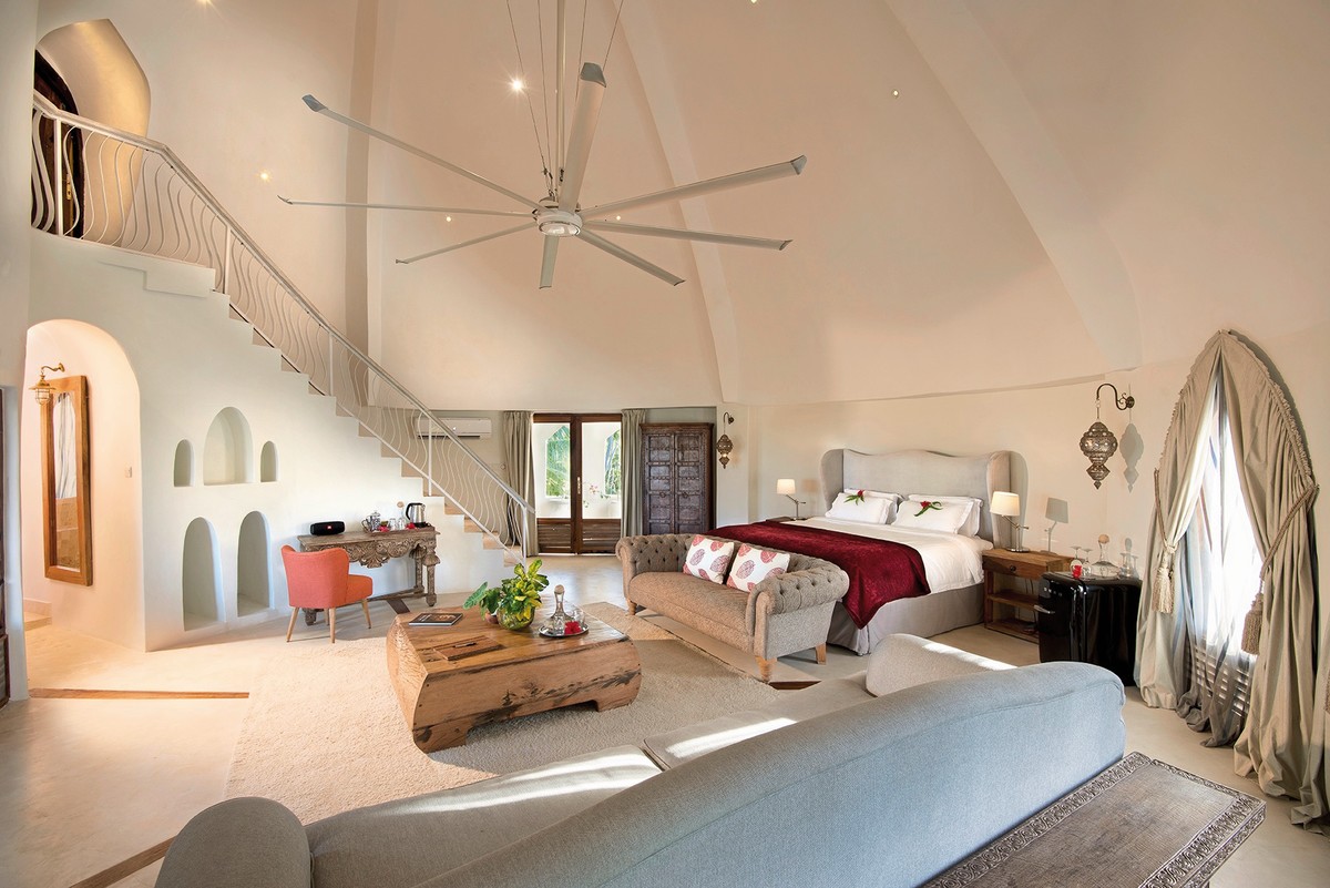 Einladendes Schlafzimmer in einem großzügigen Raum mit hohen Decken und einer elegant geschwungenen Treppe, die zu einer oberen Etage führt. Das Zimmer verfügt über ein großes Doppelbett mit weinroter Bettdecke, einen Sessel, einen rustikalen Holztisch und eine entspannende Sitzecke. Natürliches Licht flutet durch die französischen Fenster, während ein großer Deckenventilator an der Decke hängt.