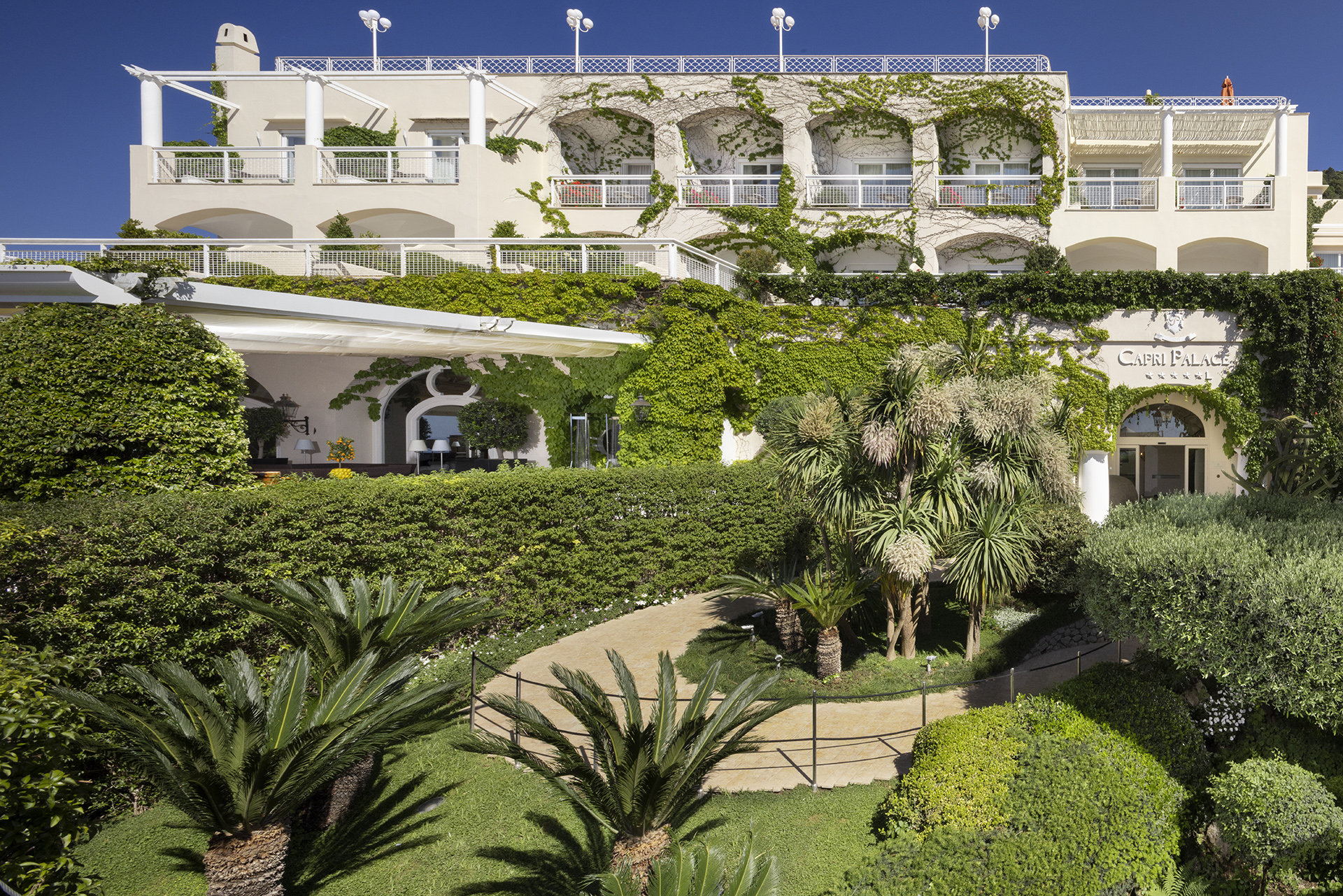 Ein sonnendurchflutetes Bild eines luxuriösen, von üppigem Grün umgebenen Hotels namens Capri Palace. Mehrstöckige Architektur mit weißen Fassaden und Balkonen, die mit Kletterpflanzen bewachsen sind. Im Vordergrund ein sorgfältig angelegter Garten mit Palmen und geschwungenen Wegen.
