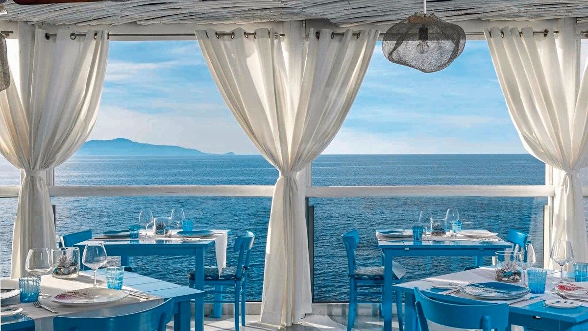Einladendes, stilvolles Freiluftrestaurant mit weiß-blauem Dekor, bietet einen atemberaubenden Ausblick auf ein ruhiges Meer und einen klaren Himmel, umrahmt von eleganten, wehenden weißen Vorhängen.