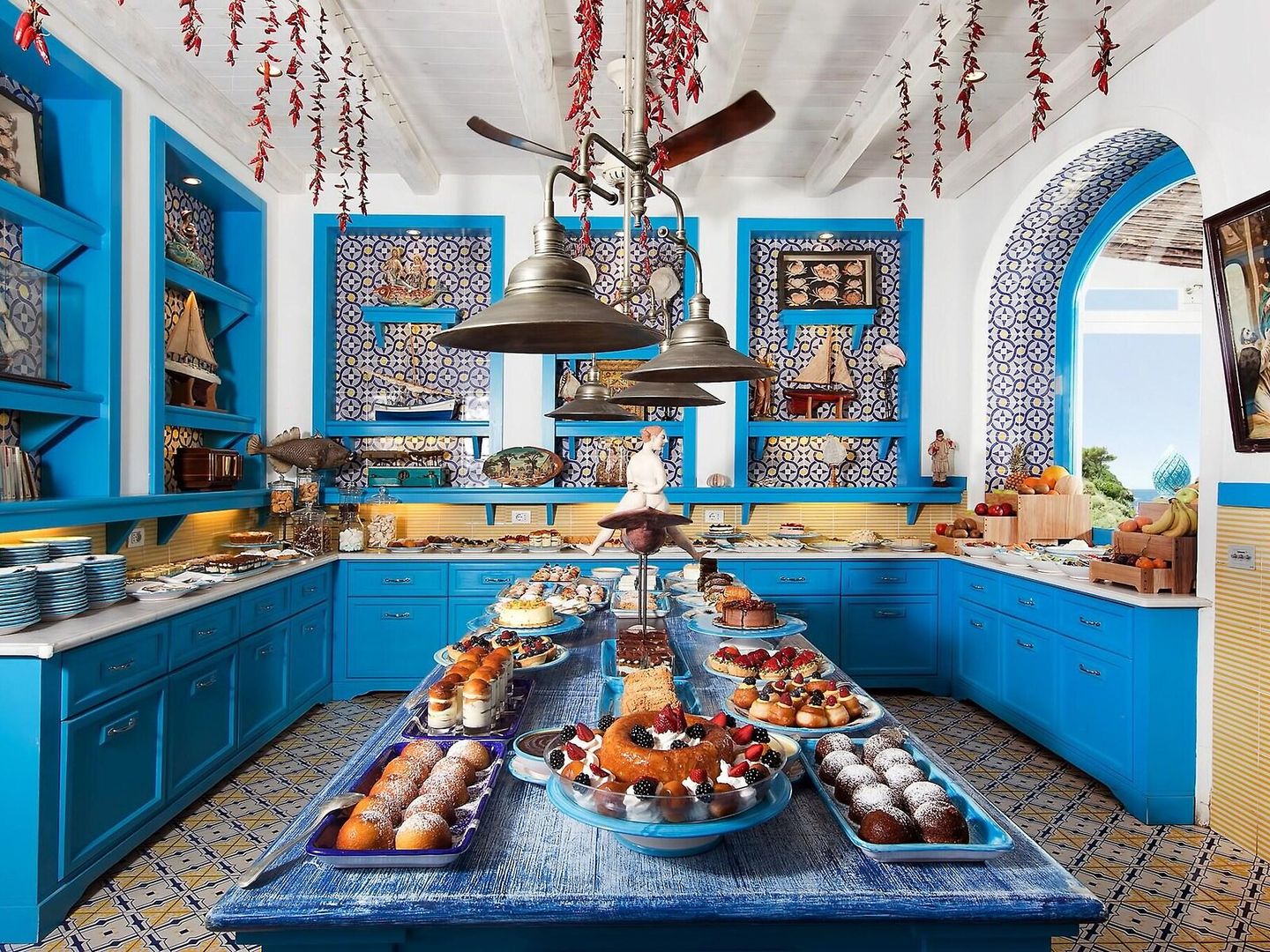 Il Riccio Restaurant Eine lebendige und farbenfrohe Küche im mediterranen Stil mit kräftig blauen Schränken und Fliesen. Eine Auswahl an Backwaren und Desserts ist kunstvoll auf dem Küchentresen präsentiert. Weiße Wände und eine bemalte Decke, kombiniert mit dekorativen Elementen, verleihen dem Raum ein einladendes Ambiente.