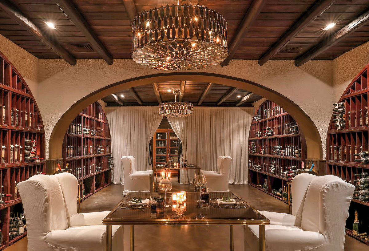 L’Olivio Restaurant Ein stilvoll eingerichtetes Esszimmer mit einem langen, reflektierenden Esstisch in der Mitte, flankiert von gepolsterten Stühlen mit weißen Hussen. Über dem Tisch hängen zwei luxuriöse Kronleuchter, die warmes Licht ausstrahlen. Der Raum wird von Holzböden und einem dunkelbraunen Deckenbalken gerahmt, und beide Seiten des Raumes sind mit hohen Regalen voll von Weinflaschen bestückt. In der Mitte des Raumes führt ein großer Bogen zu einem weiteren Raum, der durch schwere weiße Vorhänge teilweise verdeckt ist.