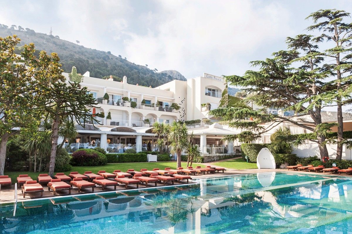 Ein luxuriöses Hotel mit weißer Fassade und mehrstöckigen Balkonen, umgeben von gepflegten Gärten und einer Reihe von Sonnenliegen neben einem großen Außenpool. Im Hintergrund erhebt sich ein bewaldeter Berg unter einem klaren, hauptsächlich wolkenlosen Himmel.