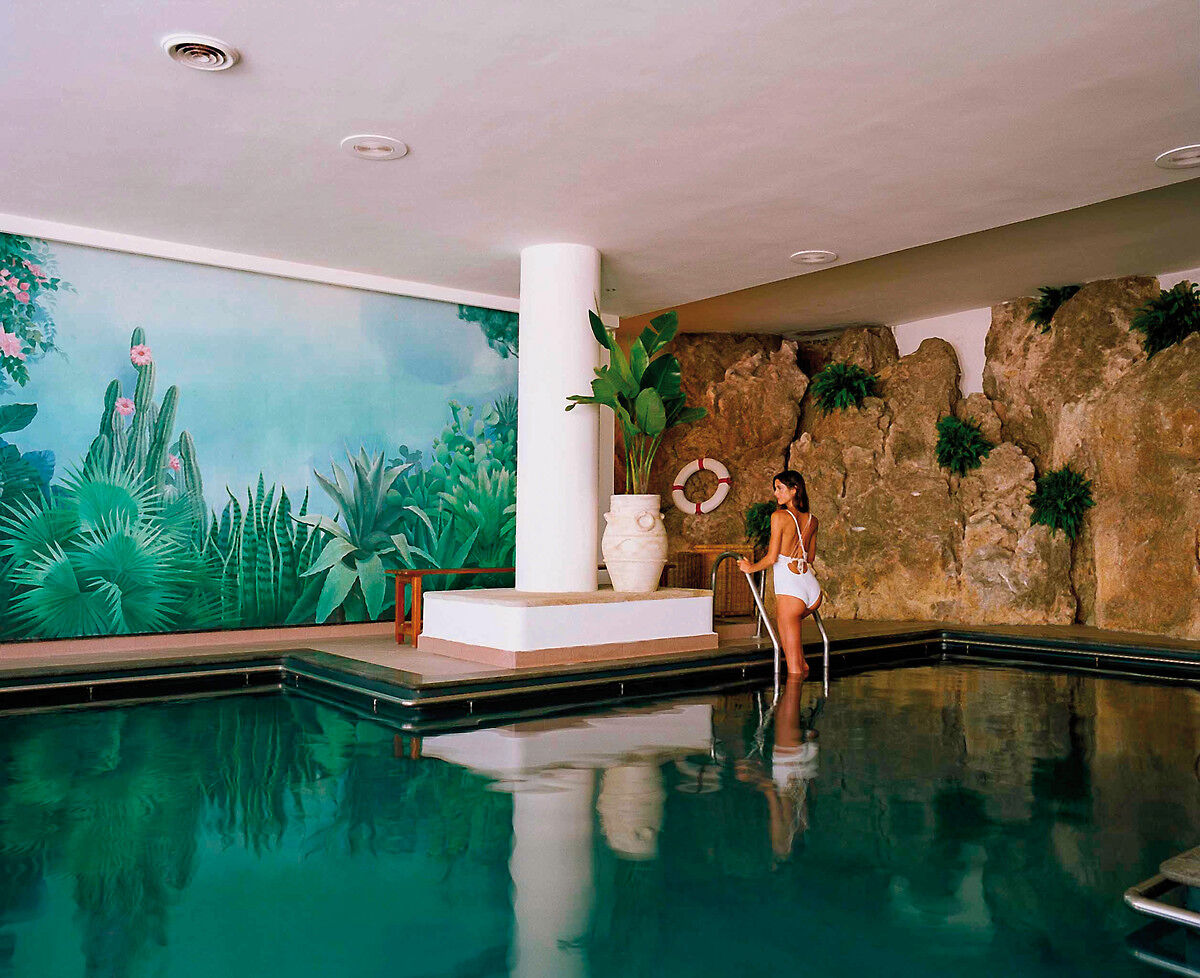 Eine Person steht am Rand eines Innenpools, der Teil eines tropisch anmutenden Wellnessbereichs zu sein scheint. Im Hintergrund ist eine Wandmalerei von tropischen Pflanzen und Kakteen zu sehen, welche die Illusion einer Fortsetzung der Landschaft ins Freie erweckt. Natürliches Licht durchflutet den Raum und spiegelt sich im ruhigen Wasser des Pools. Die Person trägt Badebekleidung und schaut entspannt in die Ferne, was eine Atmosphäre der Ruhe und Entspannung vermittelt.