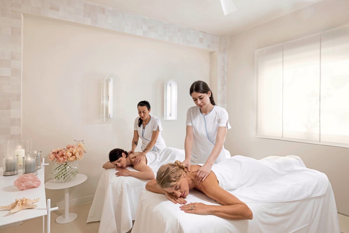 Zwei Massagetherapeutinnen in weißen Uniformen führen entspannende Behandlungen an zwei liegenden Kunden in einem hellen, modern gestalteten Spa-Raum durch. Der Raum ist schlicht mit weißen Wänden, einer großen Fensterfront und dekorativen Elementen wie Kerzen, Blumen und einem Seestern ausgestattet, die eine ruhige und friedliche Atmosphäre schaffen.