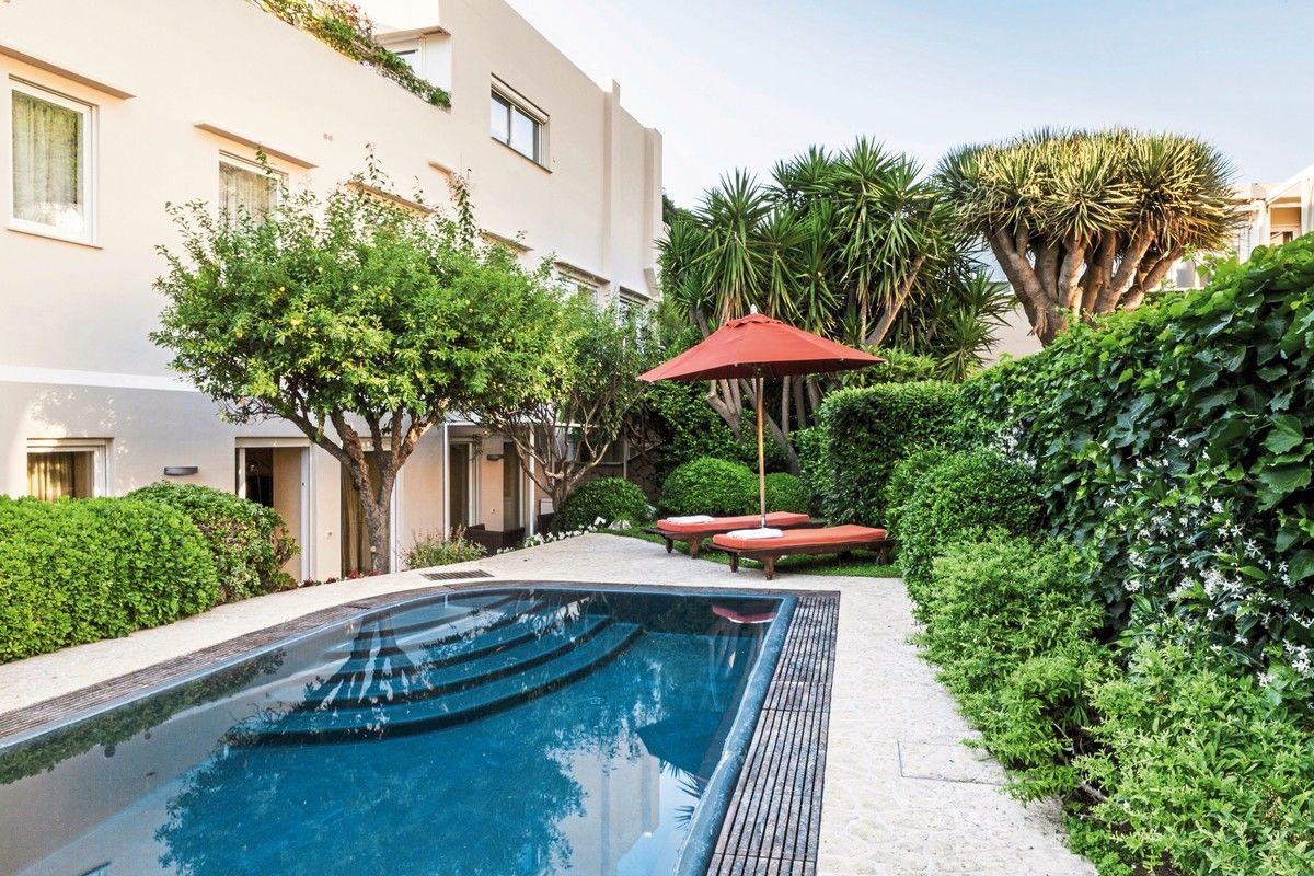 Ein modernes, weißes Wohngebäude mit Fenstern, das von einem üppigen Garten mit grünen Büschen, einem gepflegten Baum und Palmen umgeben ist. Im Vordergrund befindet sich ein einladender Swimmingpool mit klarem blauem Wasser, flankiert von einer gepflasterten Gehfläche und einer Dunkelblauen Poolleiter. Ein roter Sonnenschirm mit zwei passenden Liegestühlen unterstreicht die entspannte Atmosphäre des Außenbereichs.