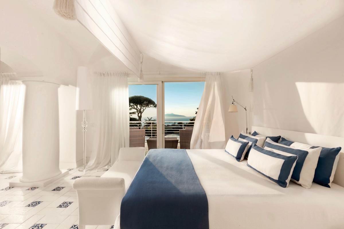 Ein helles und luftiges Schlafzimmer mit einer weißen Farbpalette, großen Fenstern und Vorhängen, durch die ein Blick auf das Meer und einige Bäume zu sehen ist. Das Zimmer ist mit einem großen Bett mit blau-weißen Kissen, einem Sofa und einem Hocker ausgestattet, und führt zu einer Terrasse mit einem Sitzbereich im Freien.