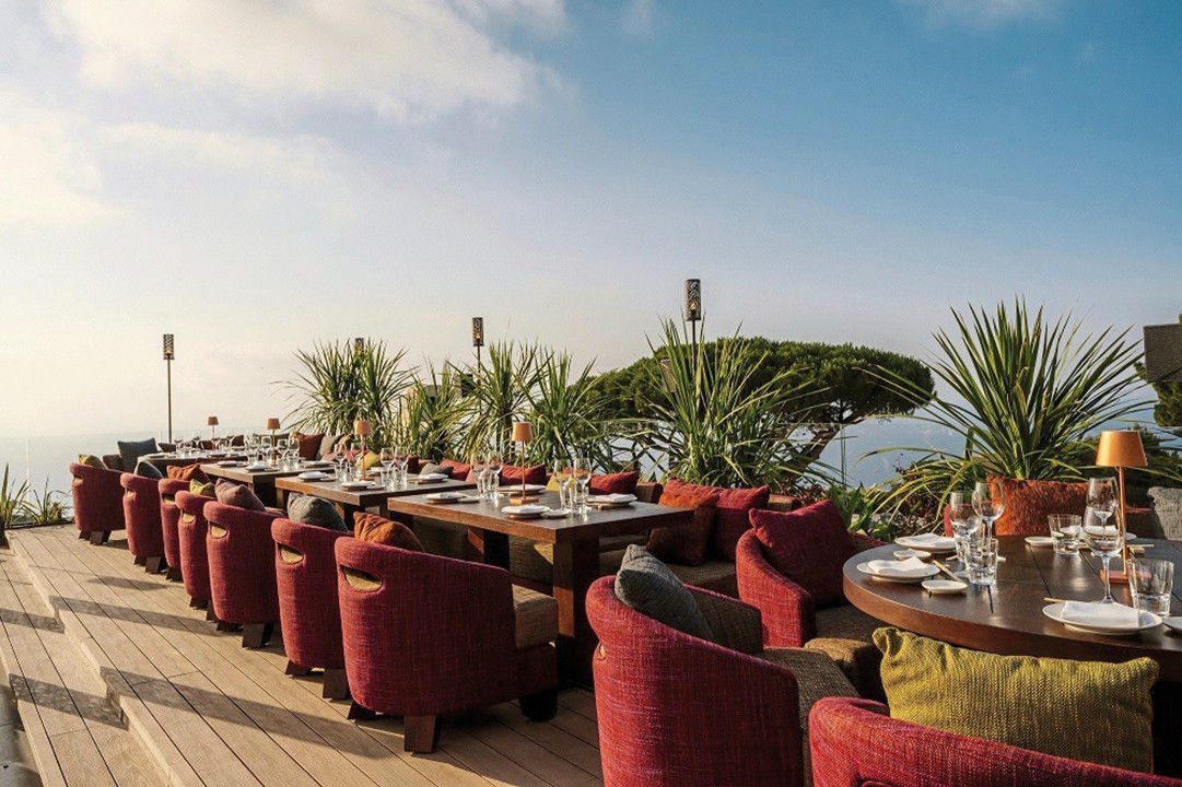 Außenterrasse eines Luxusrestaurants mit elegant gedeckten Holztischen und bequemen roten Stühlen. Grüne Pflanzen und klarer Himmel vermitteln eine ruhige Atmosphäre mit Aussicht auf das Meer.