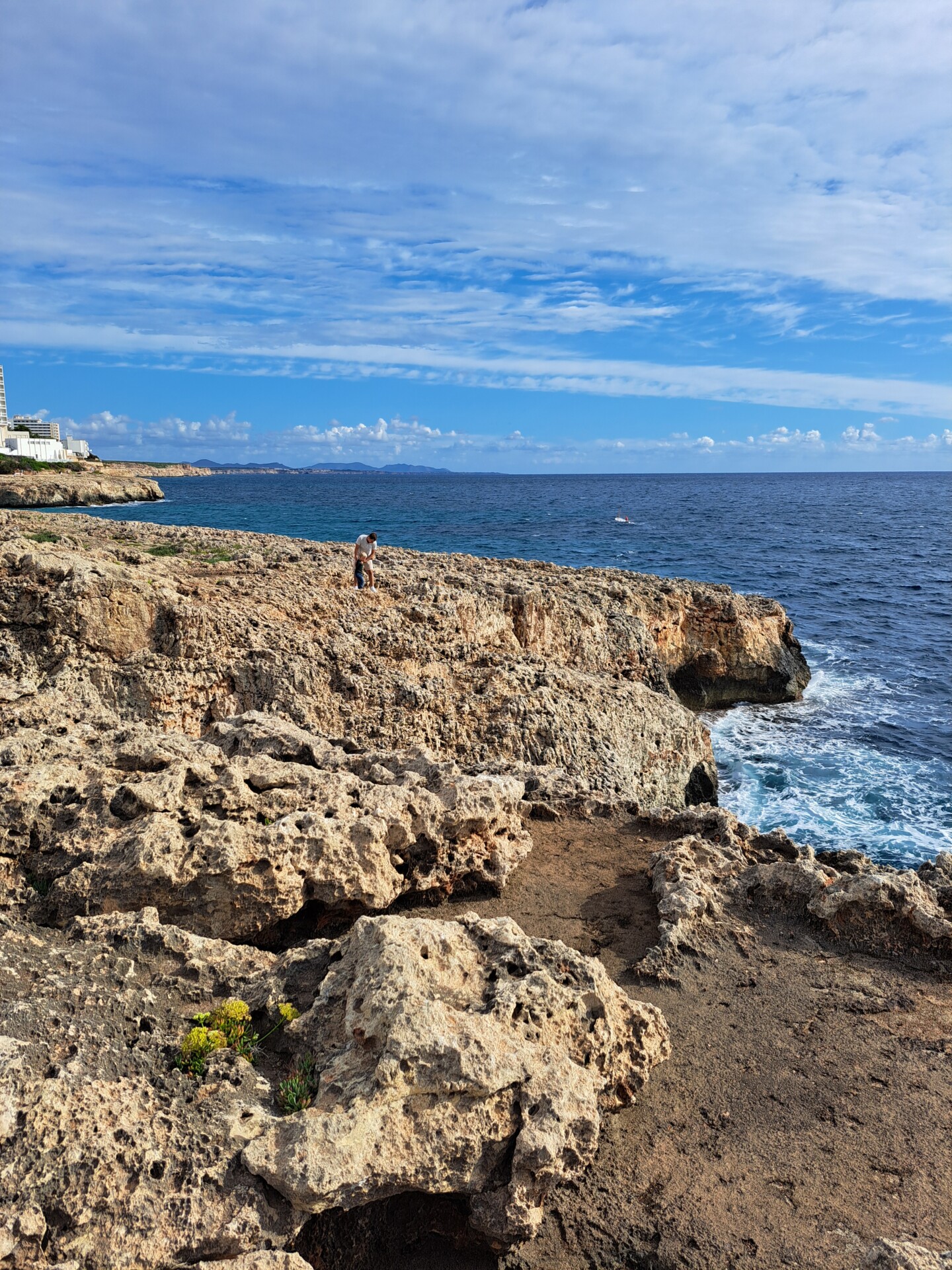 Klippen in Cala Domingos Blick auf eine raue Felsküste mit einer einzelnen Person, die auf dem Gestein steht, das Meer betrachtend. Hinter der Person liegt das ruhige, tiefblaue Meer und am Horizont ist der klare Himmel mit einigen dekorativen Wolken zu sehen.