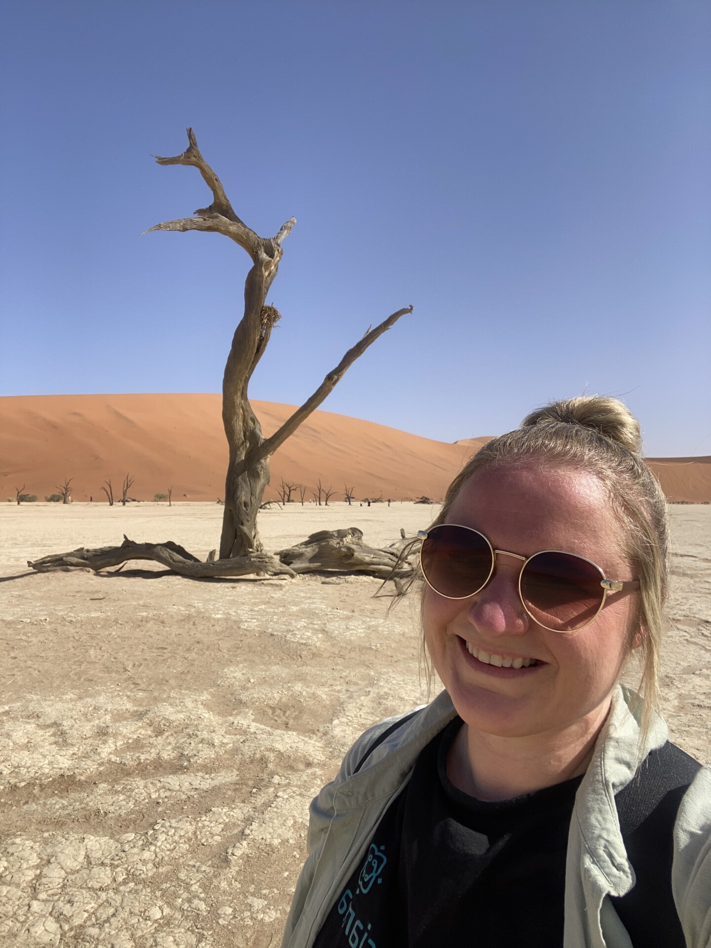 Franziska Martin im Dead Vlei Eine Frau mit sonnenbrille lächelt in der Kamera, während sie ein Selfie vor einer beeindruckenden Wüstenlandschaft macht, gekennzeichnet durch abgestorbene Bäume und große Sanddünen im Hintergrund. Der Himmel ist klar und blau.