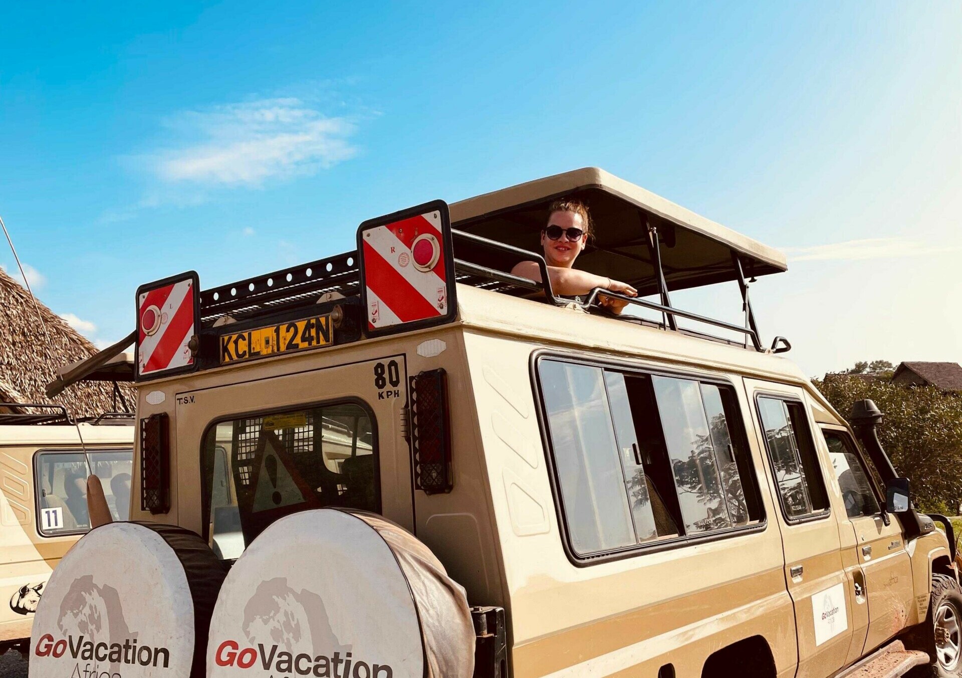 Jacqueline Fritz im Safari-Jeep Ein Frau blickt aus dem Dachfenster eines gelben Safari-Fahrzeugs heraus, das mit dem Logo von GoVacation Africa und Abbildungen eines Nashorns auf der Seite geschmückt ist. Das Fahrzeug ist mit zwei Reserverädern ausgestattet, die ebenfalls das Unternehmenslogo tragen. Die Aufnahme zeigt einen klaren blauen Himmel und eine sonnige, staubige Umgebung, die für eine Safari typisch ist.